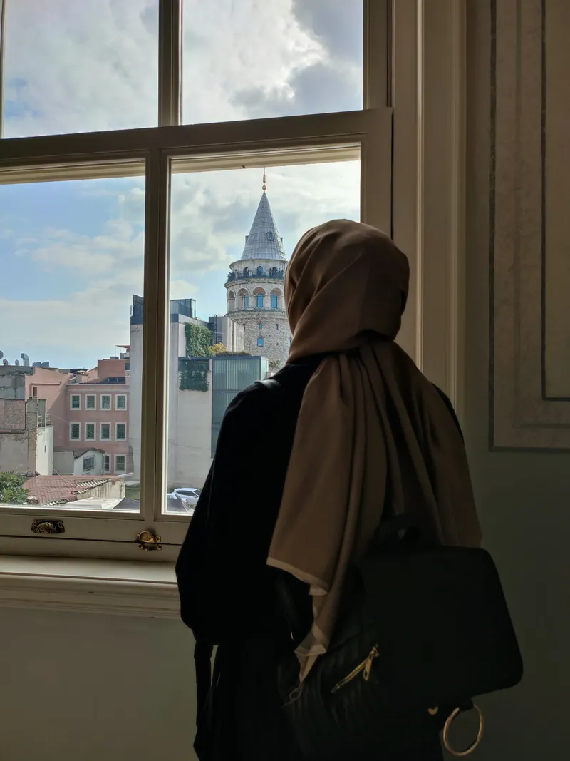 Kahverengi şal ve siyah kaban giyen kadın, pencereden Galata Kulesi'ne bakıyor